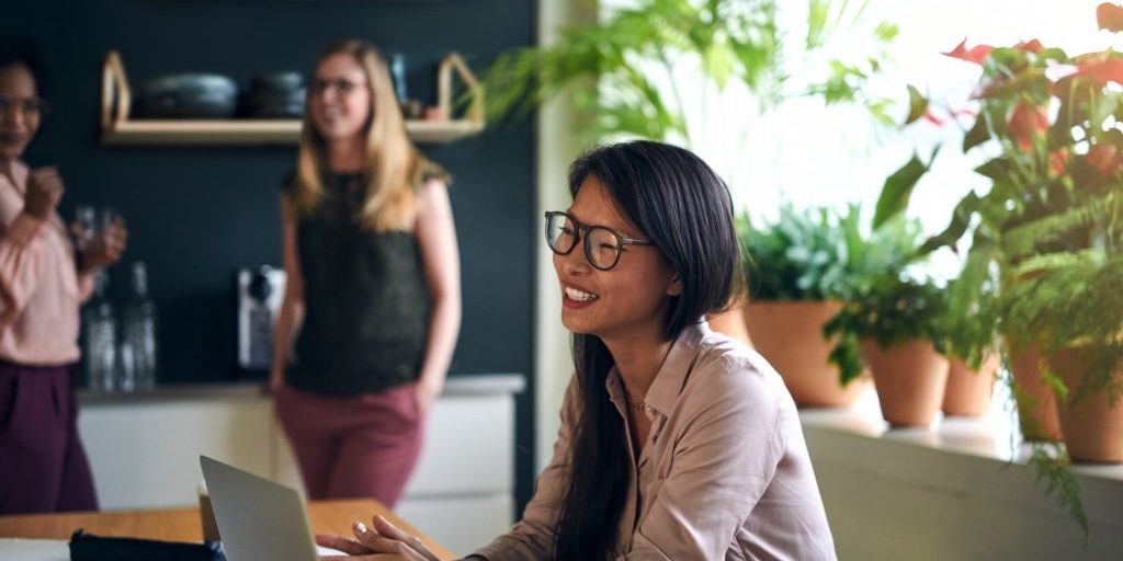 Smiling Asian businesswoman sitting at a table in a modern office using a laptop with colleagues working in the background